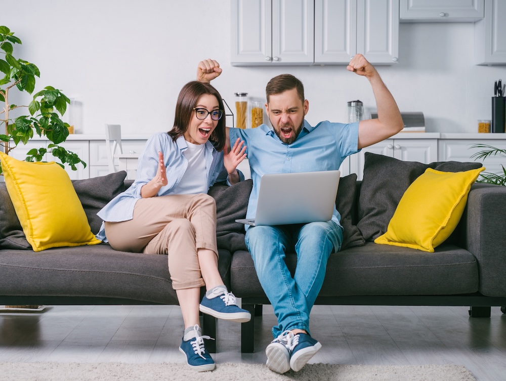 Young couple excited over results sitting on sofa with laptop, joyful